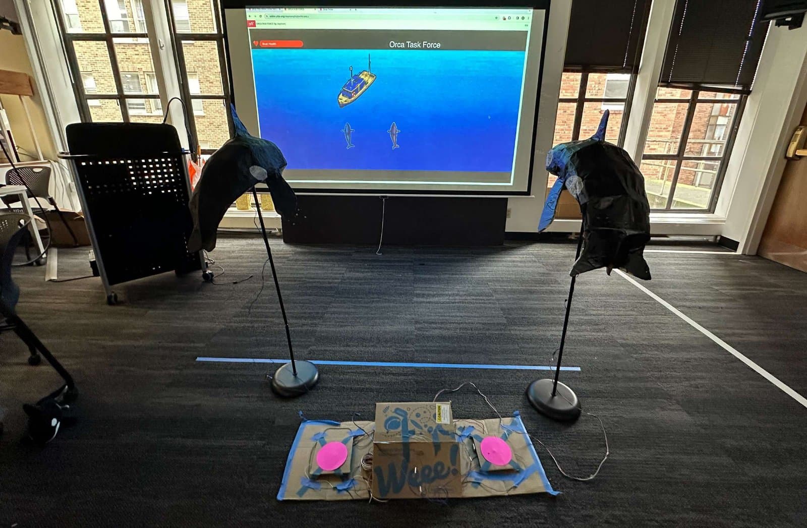 Two large cardboard orca controllers in front of a projected screen